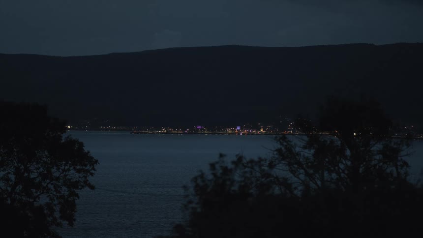 A serene view of a city's twinkling lights across a tranquil bay at night, with dark mountains in the background