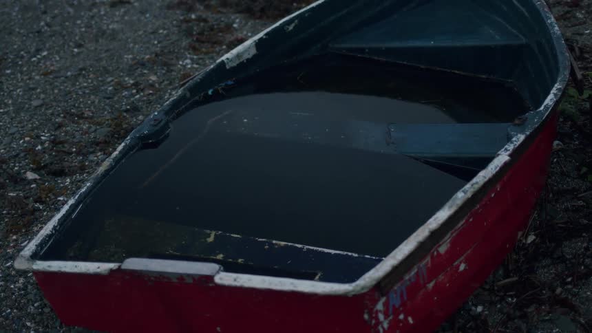 An old, red rowboat filled with dark rainwater sits abandoned on a pebbly shore