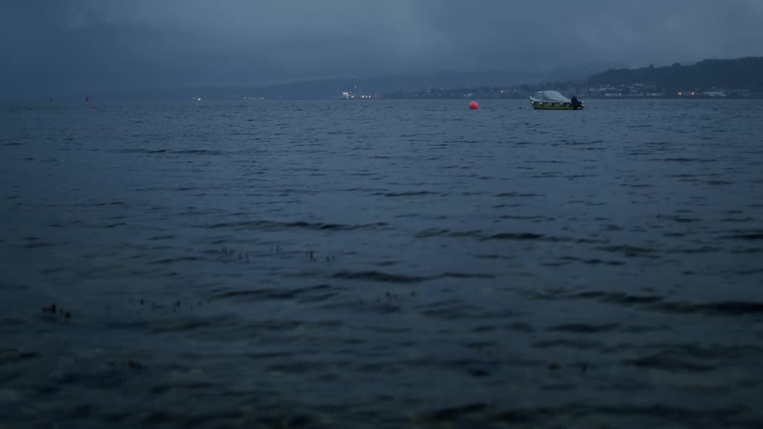Calm, dark water ripples on a lake during a gloomy evening with a lone boat anchored in the distance
