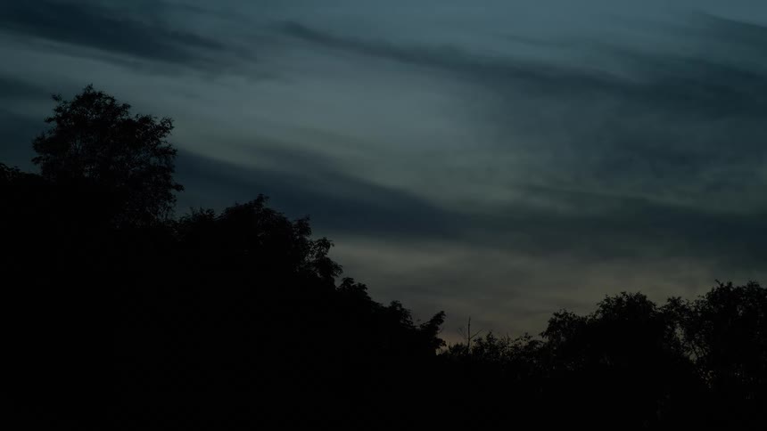 A serene, dark sky at dusk with wispy clouds moving over silhouetted trees