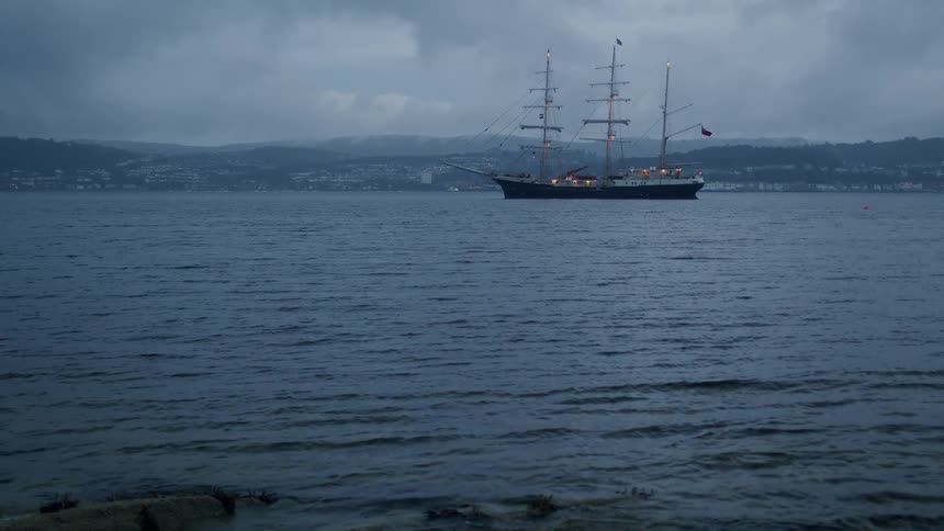 A majestic tall ship is anchored in the bay during a moody, overcast evening as waves gently ripple ashore