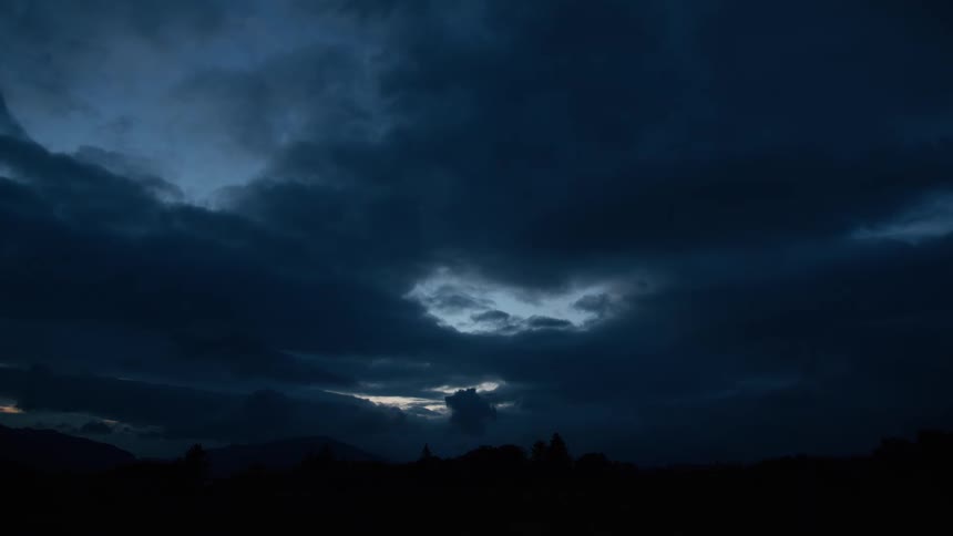 A small break in the dark, heavy clouds reveals a glimpse of light in the twilight sky over a silhouetted landscape