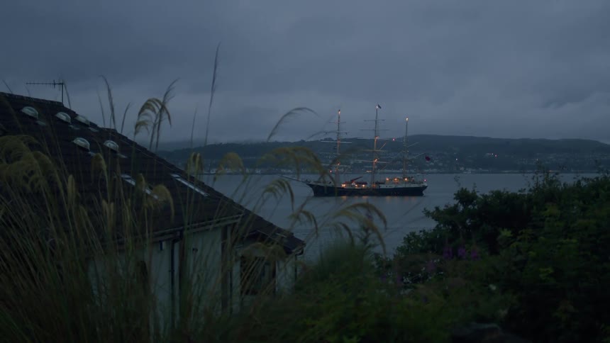 An illuminated tall ship is anchored in a bay at dusk, viewed from a quiet, grassy hillside