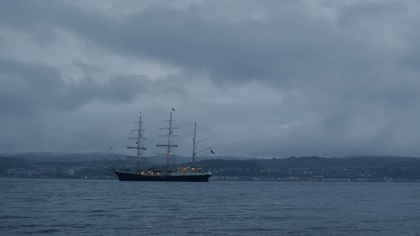 A historic tall sailing ship is anchored in the bay on a moody, overcast evening