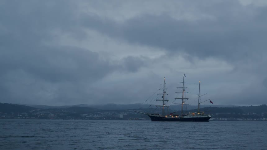 A majestic tall ship floats on calm waters near a distant shoreline on a gloomy, overcast evening