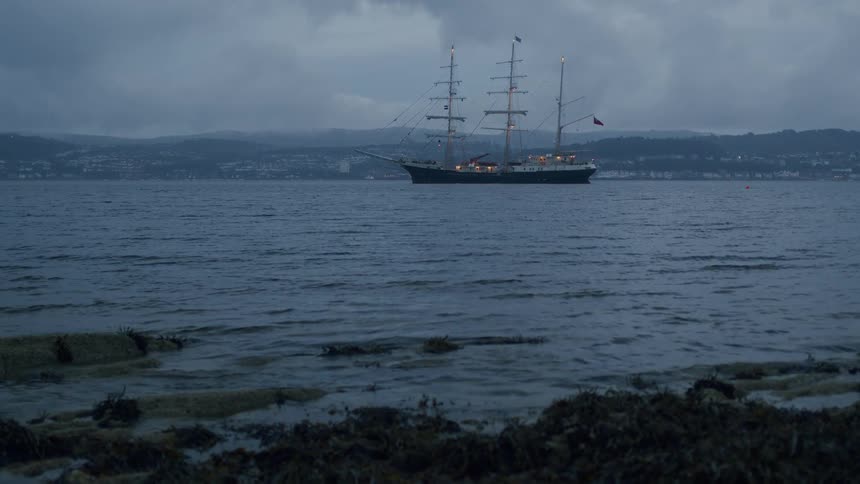 A majestic tall ship is anchored in a calm bay on a cloudy evening as gentle waves wash over the seaweed-covered shore