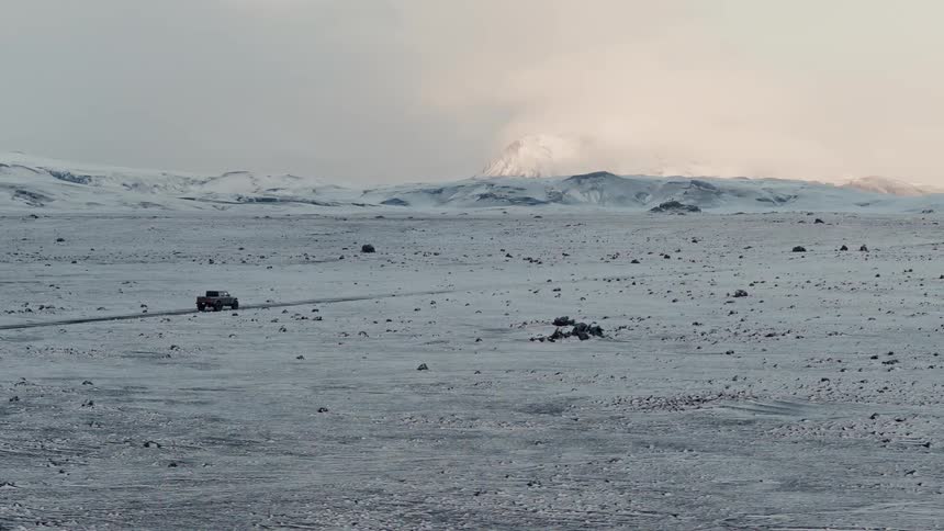 A rugged truck drives along a remote road through a vast, snow-covered landscape with mountains on the horizon