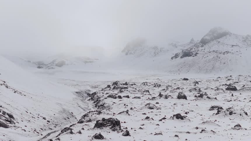 A slow zoom-out of a desolate, snow-covered, and rocky valley with foggy mountains in the background