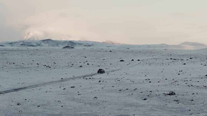 A wide shot shows a single off-road vehicle driving through a vast, desolate, snow-covered landscape in Iceland with mountains in the background
