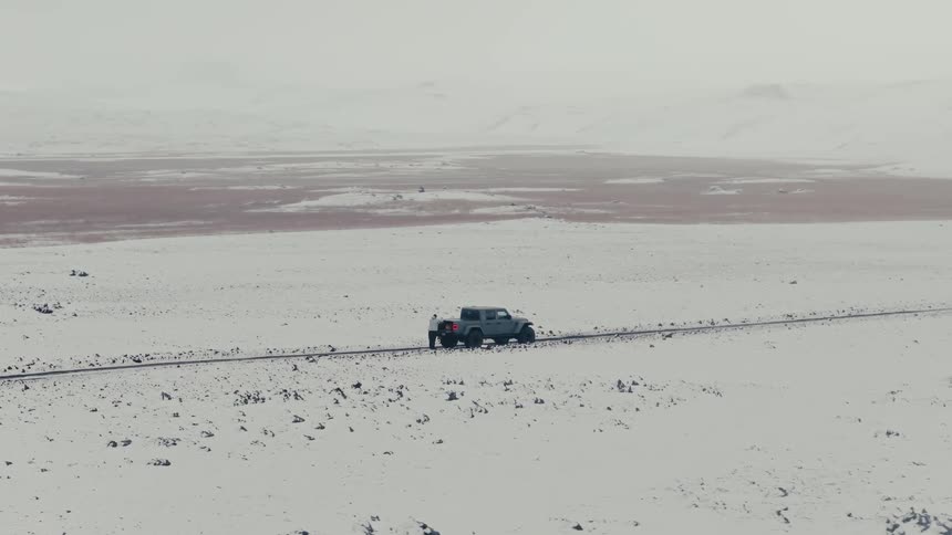 An aerial view shows a person getting into a pickup truck and driving down a remote road through a vast, snow-covered landscape