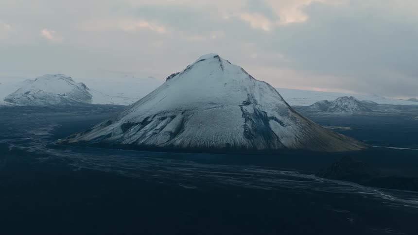 A static wide shot captures the dramatic, desolate landscape of a snow-capped volcanic mountain in Iceland under an overcast sky
