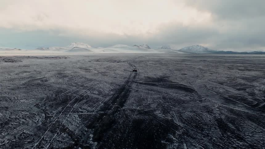 An aerial shot follows a lone truck driving through a vast, desolate, snow-covered landscape towards distant mountains