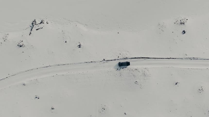 Aerial top-down view of a single car driving on a snowy road through a vast, empty winter landscape