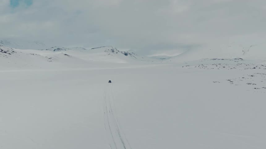 An aerial view captures a solitary vehicle driving across a vast, endless snow-covered landscape towards distant mountains