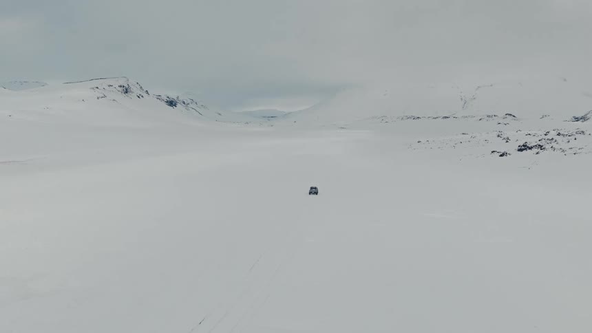 An aerial view captures a solitary vehicle driving through a vast, desolate, snow-covered landscape with mountains in the background