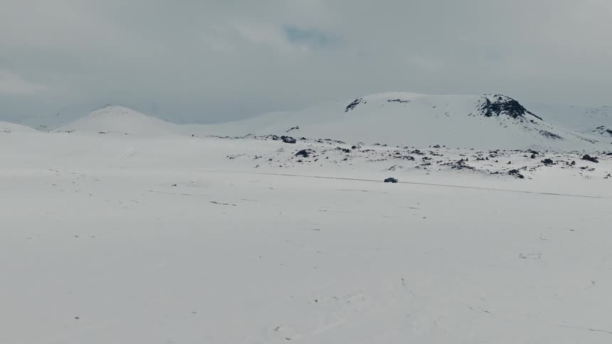 An epic wide aerial shot captures a lone car driving through a vast and desolate snowy mountain landscape under a cloudy sky