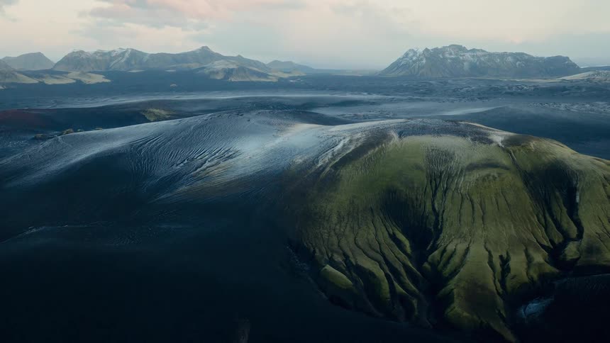 A breathtaking, static aerial view captures the otherworldly beauty of Iceland's volcanic highlands, with mossy green hills and snow-capped mountains