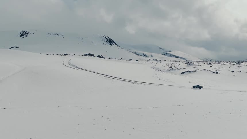 An aerial shot follows a lone truck as it drives across a vast, snow-covered landscape with mountains in the background