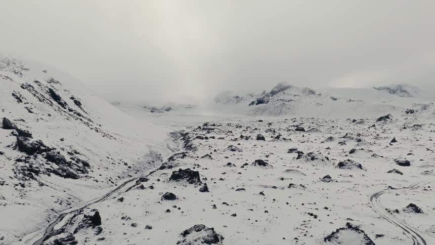 A slow aerial pullback reveals a vast, desolate valley of snow-covered mountains and rocks on a bleak winter day