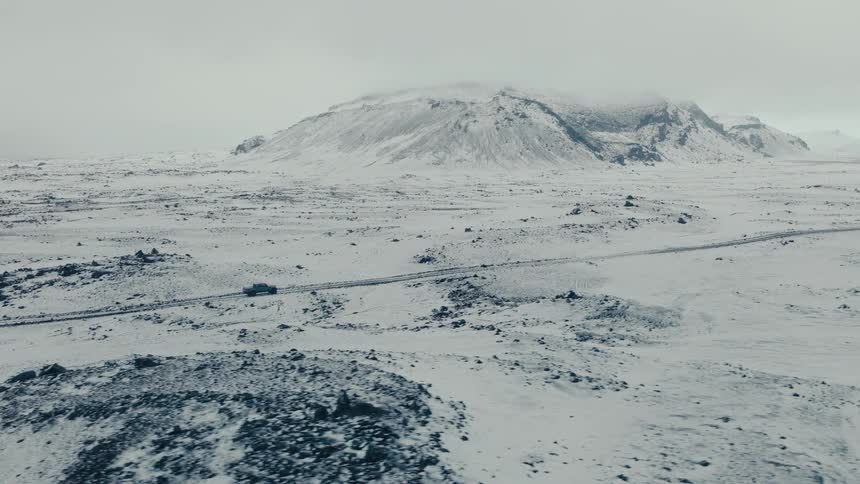 Aerial footage tracks a rugged off-road vehicle driving through an immense, desolate, and snow-covered terrain towards a distant mountain