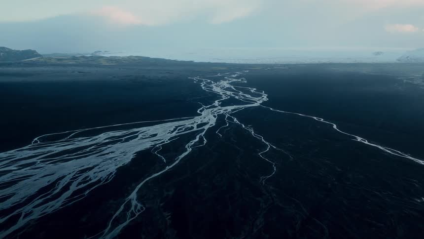 An epic aerial view of a milky white glacial river carving its path through a vast black volcanic sand plain towards a distant glacier