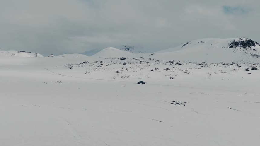 Wide aerial drone shot follows a lone 4x4 vehicle driving across a vast and desolate snow-covered mountain landscape