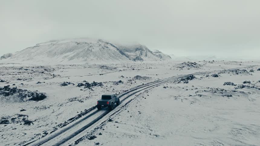 Aerial shot following a pickup truck driving on a remote dirt road through a vast, snow-covered mountain landscape