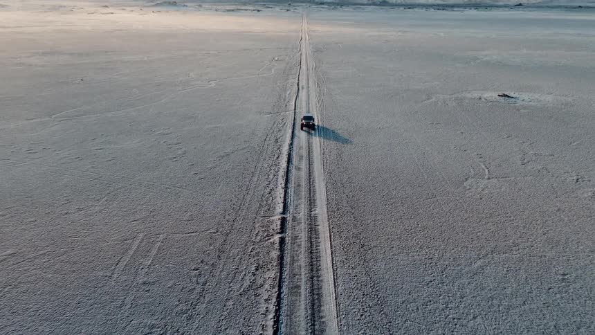 A drone follows a lone car driving through a vast, remote, snow-covered landscape with mountains on the horizon