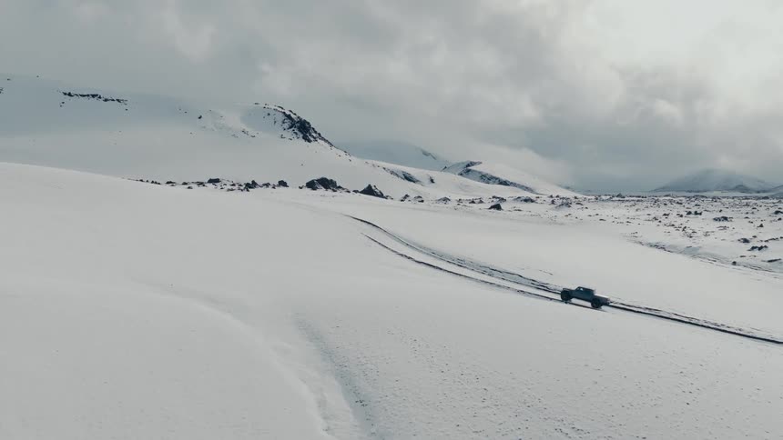An aerial view captures a large monster truck driving through a vast and desolate snow-covered landscape