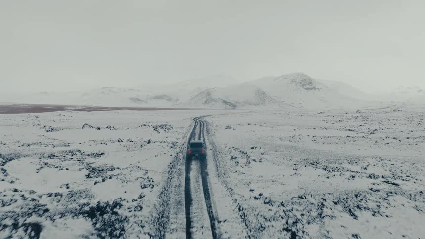 An aerial view follows a lone SUV as it travels down a remote road through a vast, desolate, snow-covered landscape towards distant mountains