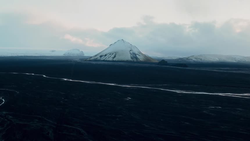 A majestic snow-capped mountain rises from a vast, dark, frozen landscape under an overcast sky