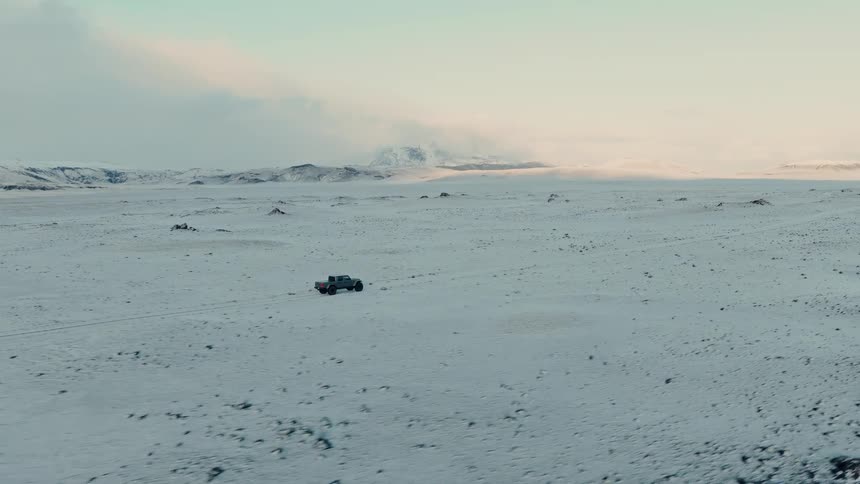 An aerial shot follows a lone pickup truck driving through a vast, desolate snow-covered landscape towards distant mountains