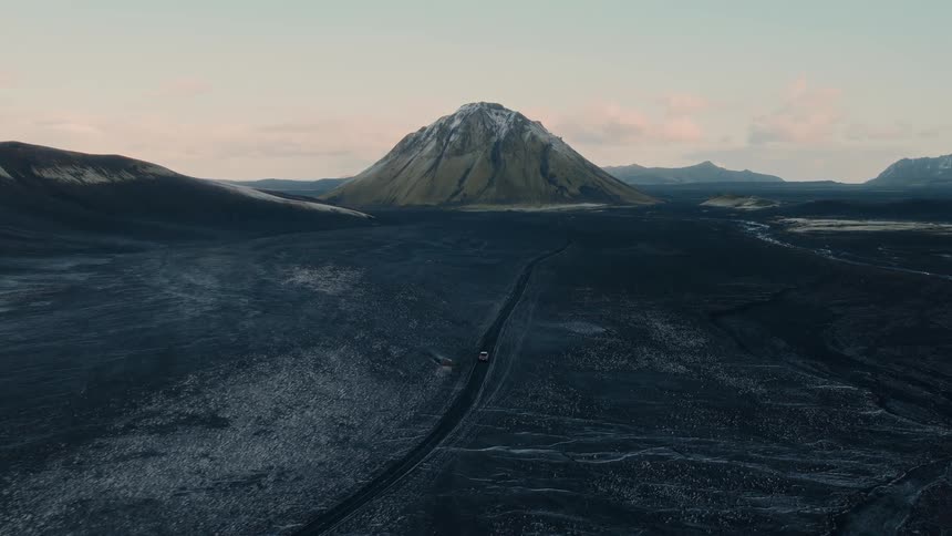 An epic aerial view of two cars driving through a vast, dark volcanic landscape towards a majestic snow-capped mountain in Iceland