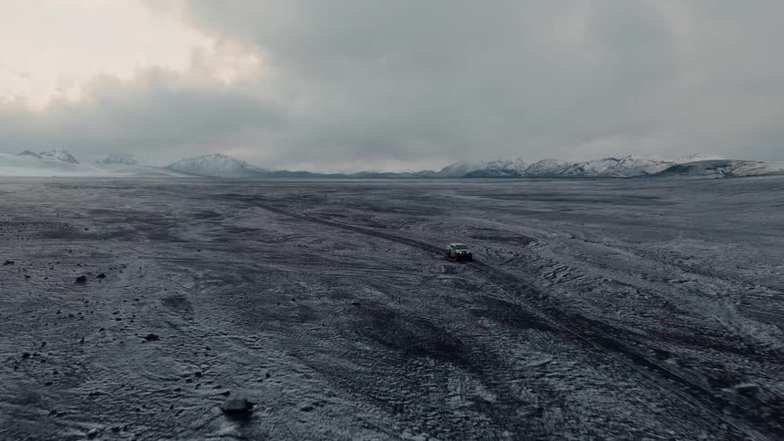 A lone SUV travels down a desolate road through a vast, epic landscape of snow-dusted volcanic plains under a cloudy sky