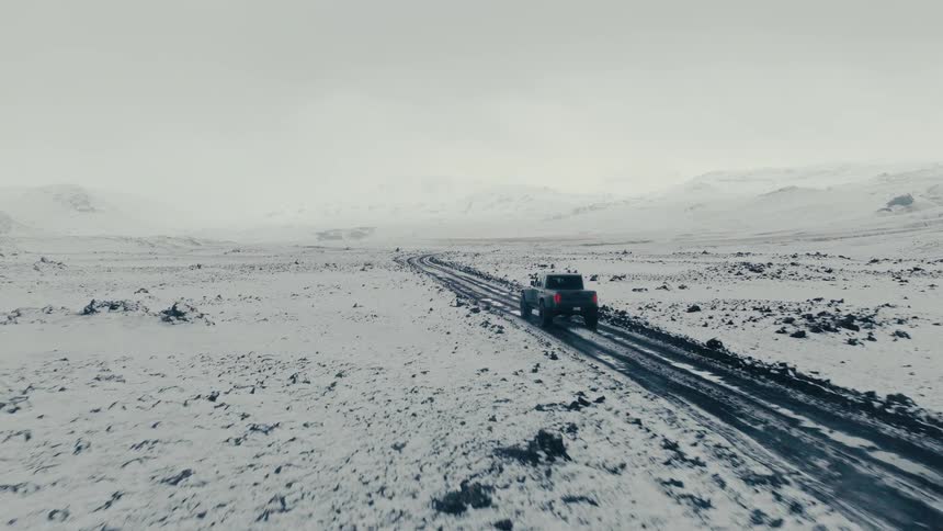 An aerial view follows a rugged pickup truck driving down a muddy road through a vast, snow-covered, and desolate landscape