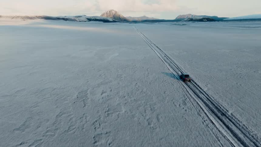 An aerial view follows a lone SUV driving through a vast, desolate, snow-covered landscape towards distant mountains