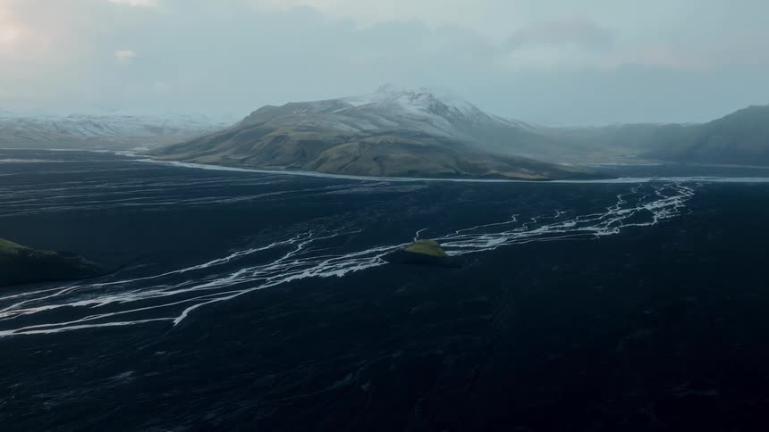 Wide aerial shot of a breathtaking Icelandic landscape shows a network of glacial rivers flowing through a vast black volcanic plain towards snow-dusted mountains