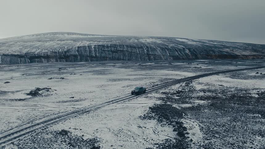 An aerial view reveals a lone SUV navigating a remote road through a vast, snow-covered, and desolate Icelandic landscape