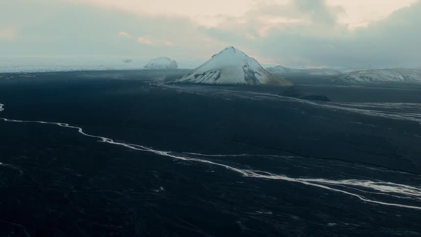 A vast and moody Icelandic landscape features a solitary snow-capped mountain rising above a dark volcanic plain and winding river