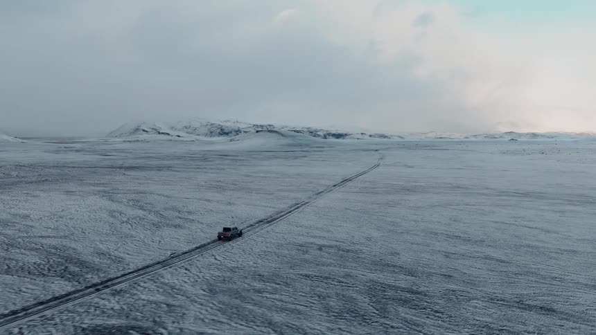 A dramatic aerial shot follows a lone off-road vehicle as it travels through a vast and desolate snow-covered landscape towards distant mountains
