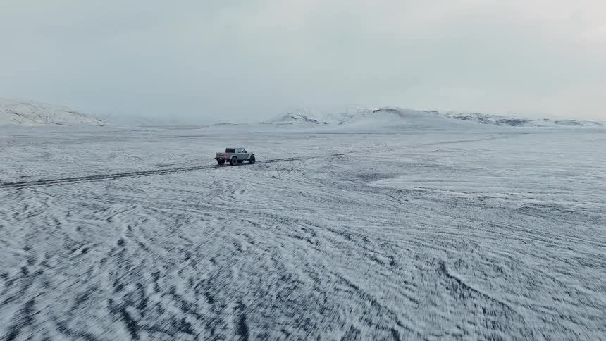 Aerial view of a large 4x4 pickup truck driving off-road through a vast and remote snow-covered landscape towards distant mountains
