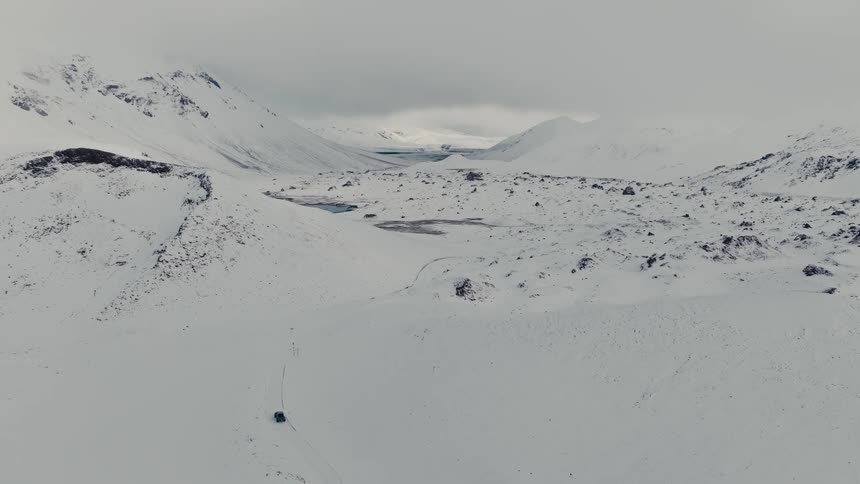 An epic aerial shot follows a lone vehicle driving through a vast and desolate snow-covered mountain valley