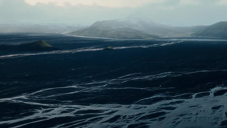 A still shot captures the vast, dark valley with silver streams of a braided river flowing towards misty, snow-capped mountains