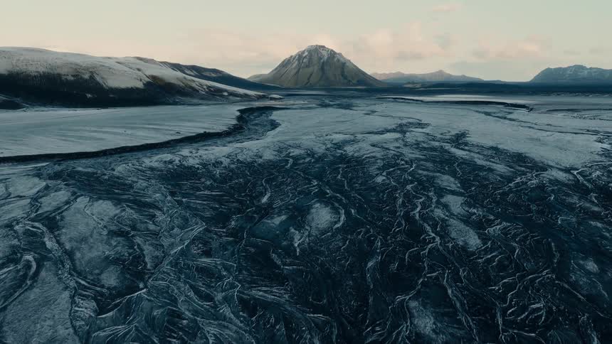 A slow aerial tilt-up reveals the epic, desolate beauty of a frozen braided river and a snow-dusted mountain in a vast Icelandic landscape