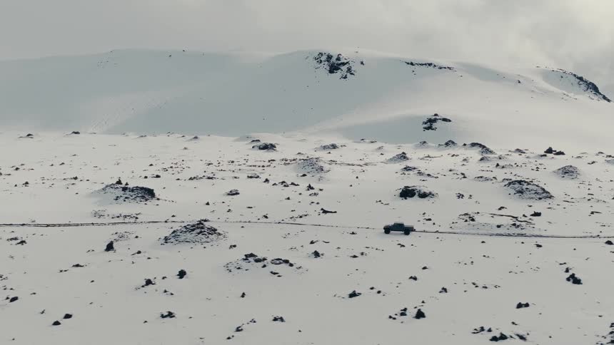 An aerial view captures a pickup truck driving along a remote road through a desolate, snow-covered mountain landscape