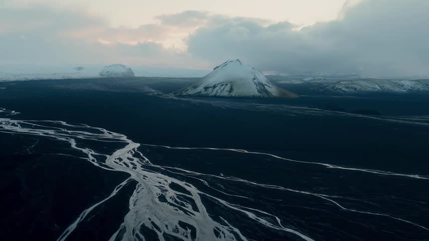 A stunning, moody aerial shot of Iceland's otherworldly landscape, featuring frozen river braids snaking through black volcanic plains towards a snow-capped mountain