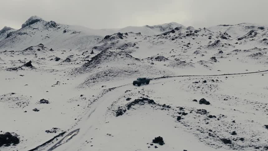 An aerial view follows an SUV driving along a remote, winding road through a vast, snow-covered mountain landscape