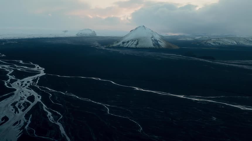 An otherworldly Icelandic landscape features a vast, dark plain with intricate patterns of a braided river system and distant snow-covered mountains at dusk