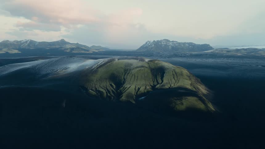 A wide, epic aerial shot captures a lone, moss-covered green mountain rising from a vast black volcanic sand desert in the Icelandic highlands