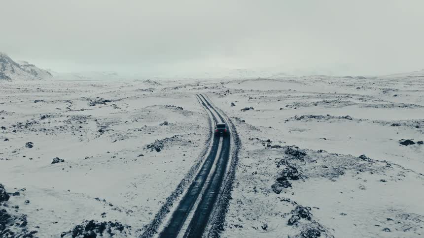 An aerial view shows a lone SUV driving down a desolate, winding road through a vast, snow-covered landscape